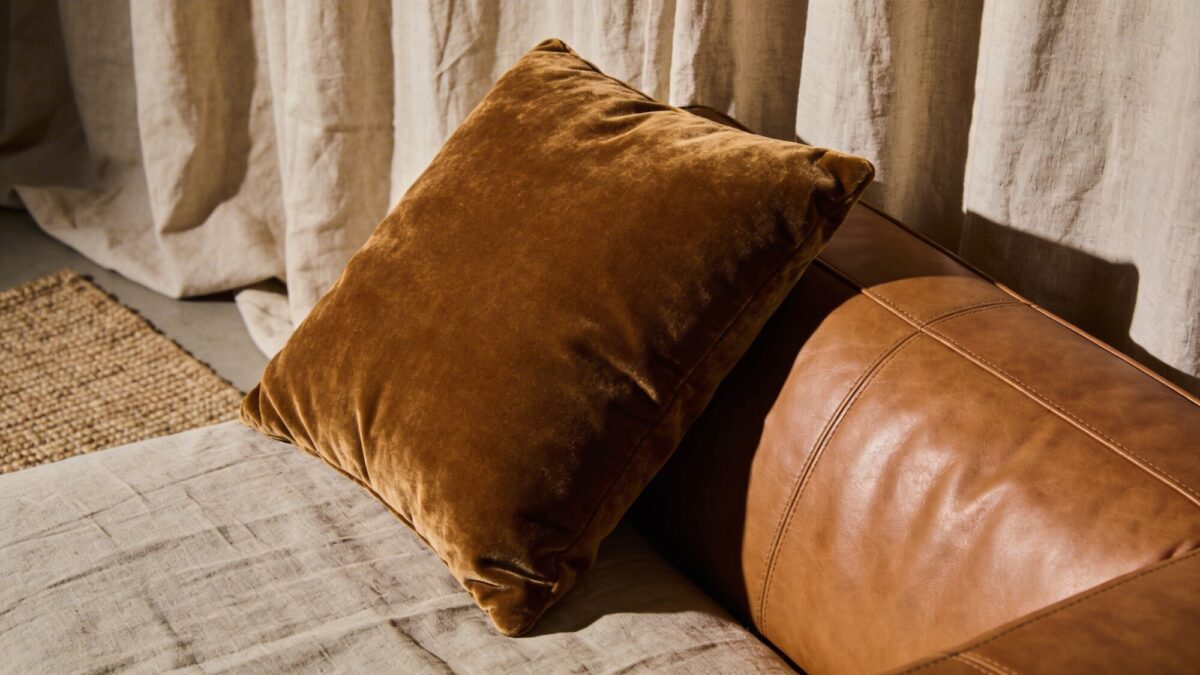 Close-up of layered textures: a velvet cushion resting on a leather sofa with a linen drape backdrop, a tactile woven rug visible on the floor, soft directional lighting highlighting the fabric contrasts.