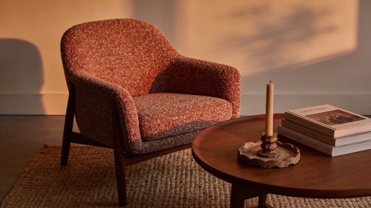 A warmly lit living room at dusk with soft ambient lighting, a boucle-textured armchair beside a round wooden coffee table, a stack of books, a candle centerpiece, and a woven rug; neutral palette with hints of warm rust.