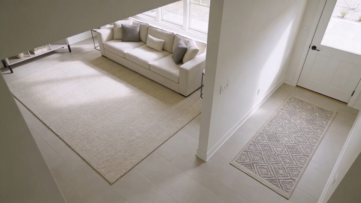 A bright, airy living room showing layered rugs: a larger base rug under the seating area with a smaller patterned runner near a doorway, demonstrating how scale defines zones without overwhelming space.