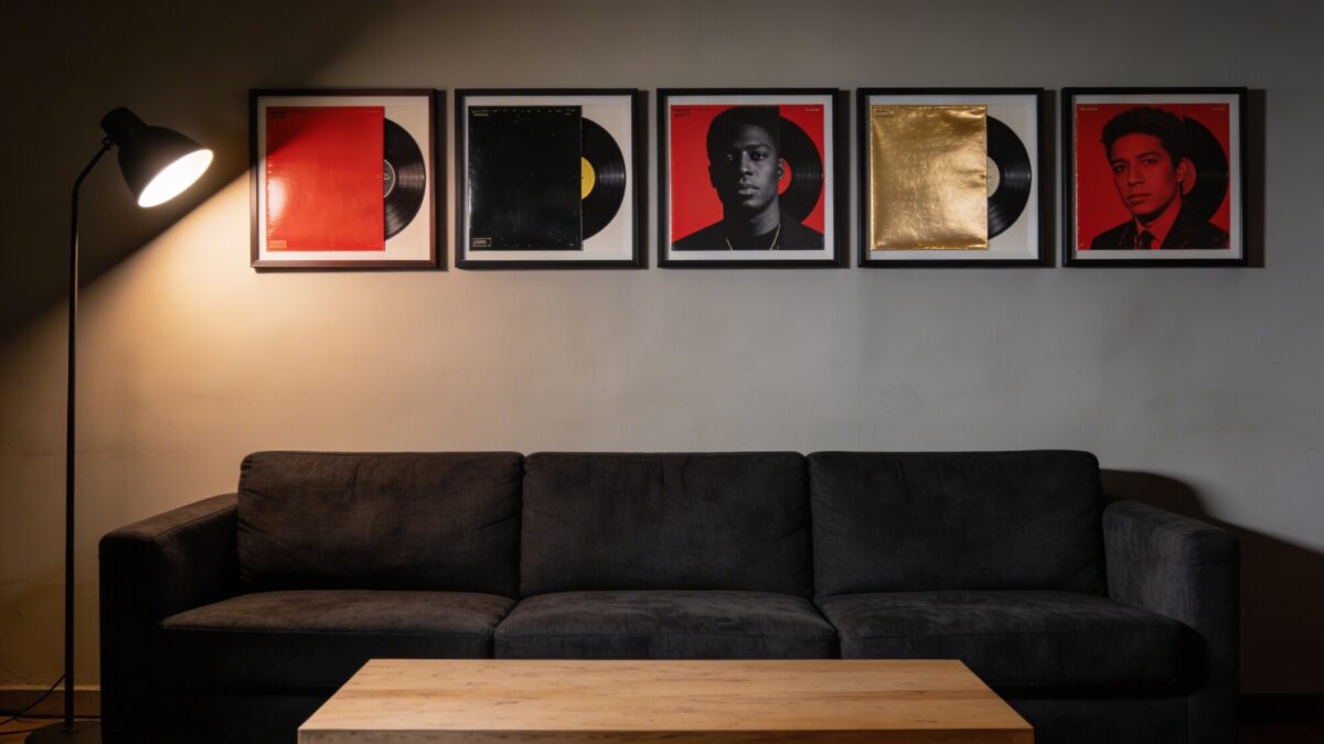 A moody, high-contrast gallery wall: five album covers with bold color blocks (red, black, gold) in exact same frame style, even spacing, dramatic side lighting from a floor lamp, a dark charcoal sofa, and a light wood coffee table to balance the composition.