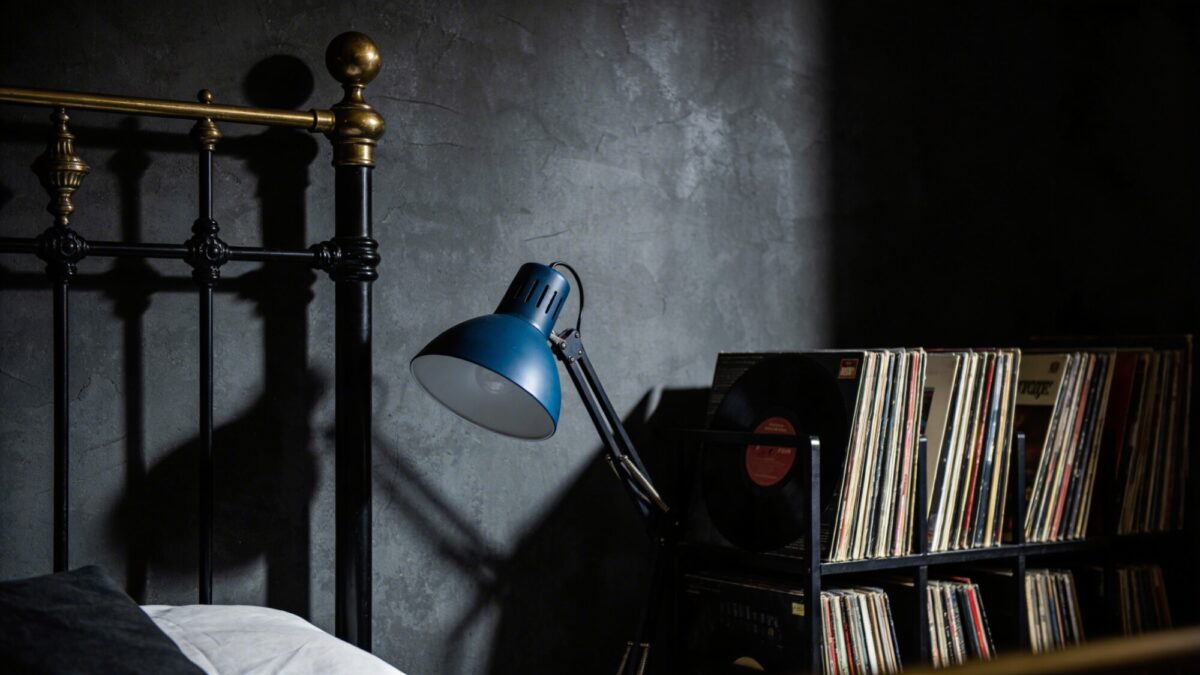 Moody noir album vibe: charcoal gray walls, a black metal bed frame, gilded brass accents, rich obsidian and slate textures, a single moody blue lamp casting dramatic shadows, and a shelf filled with vinyl records, no text.