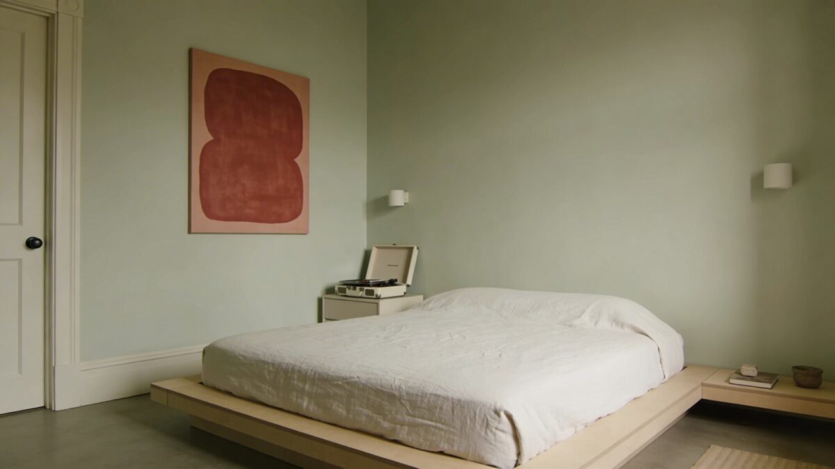Indie minimalist retreat: pale sage walls, a platform bed with a simple white duvet, a single bold abstract print in muted rust, a compact record player on a clean nightstand, and sparse but intentional decor, no text.