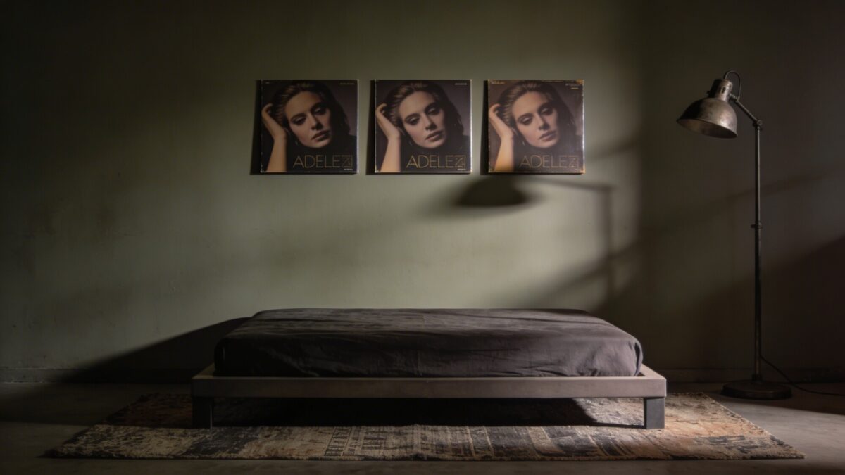 A triptych of three evenly spaced album covers arranged to evoke a moody, cinematic vibe, hung above a low-profile platform bed with a textured rug, muted wall color, and a single industrial-style floor lamp casting gentle shadows.