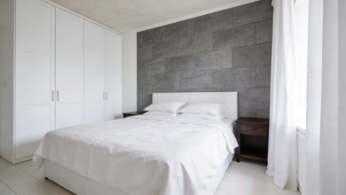 A “don’t overdo white” tip visualization: a bright bedroom with mostly white elements, but showing clean dust-free surfaces, a slightly off-white ceiling, and a contrasting slate gray wall and dark wood nightstands to illustrate balanced contrast.