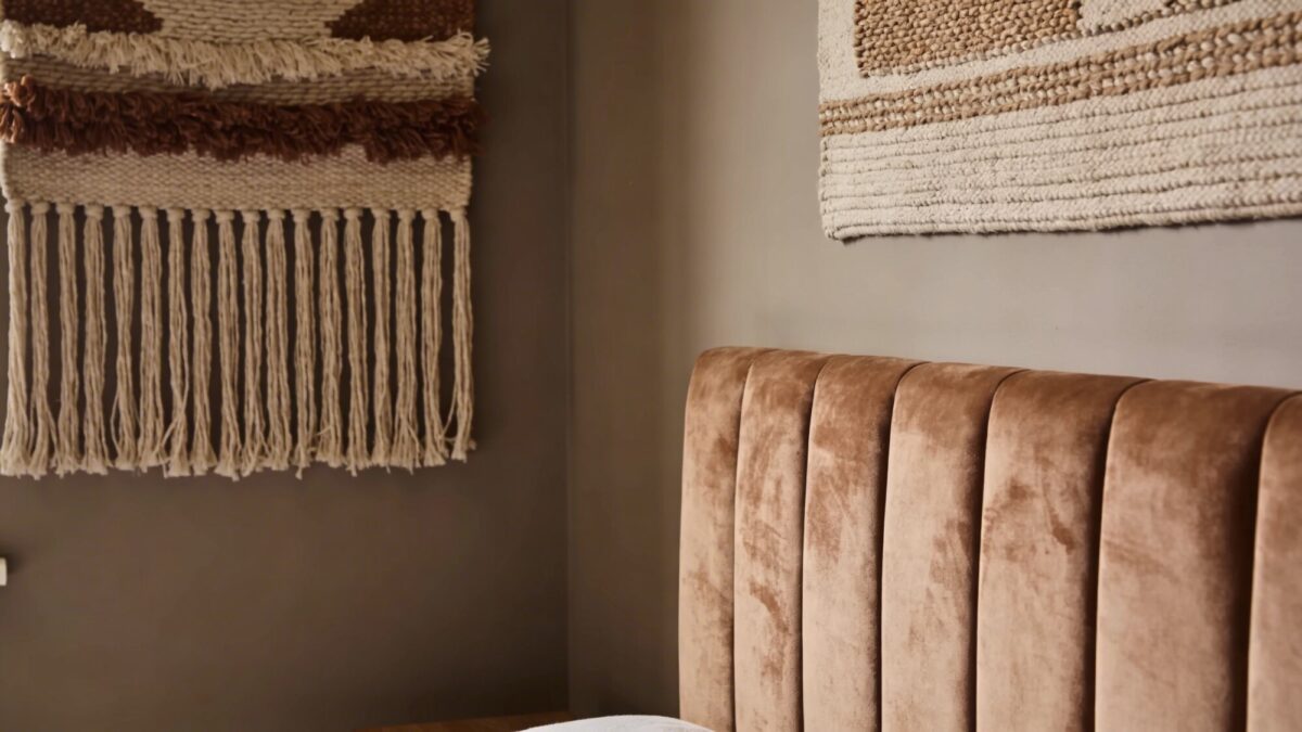 Close-up of a textured wall: a soft matte taupe wall paired with a woven tapestry and a suede headboard, emphasizing layered textures over patterns in a small room.