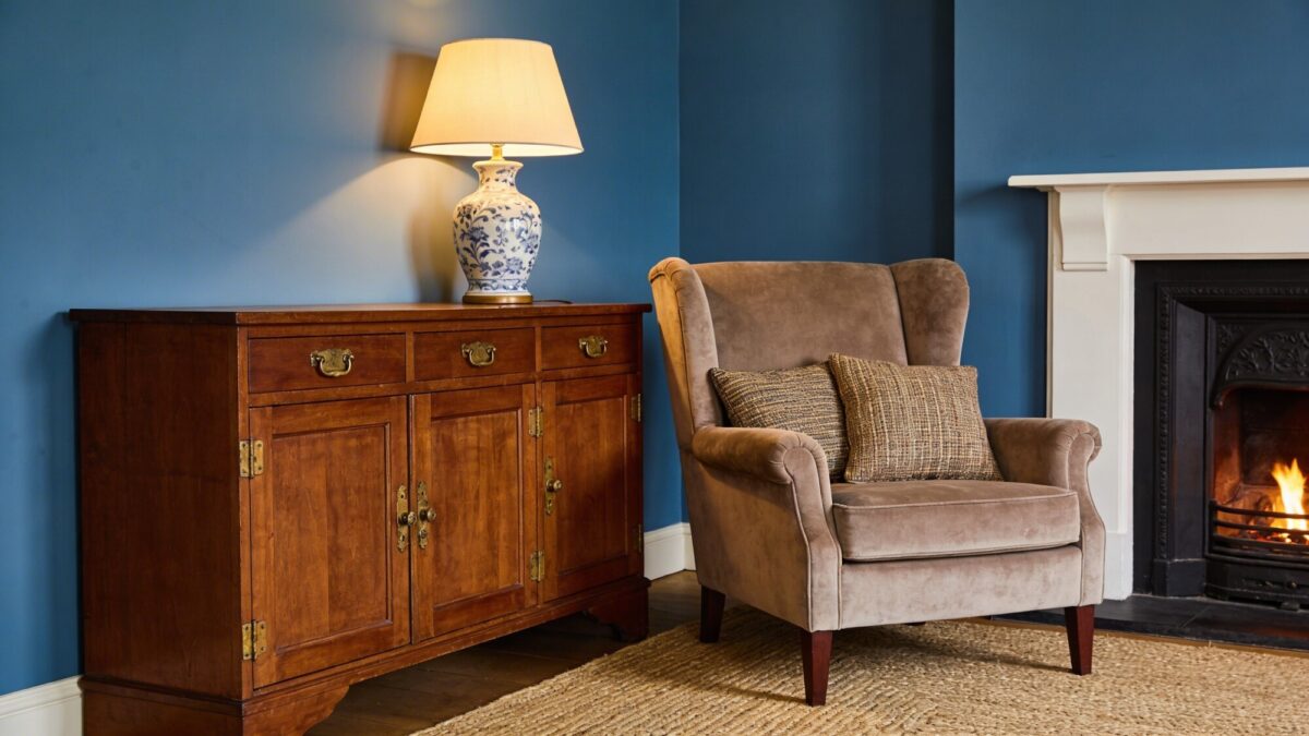 A cozy corner scene featuring a suede armchair, a rich oak sideboard with aged hardware, a porcelain lamp, and a fireplace with a simple mantel; rich textures like jute rug, tweed cushions, and a blue and cream palette.